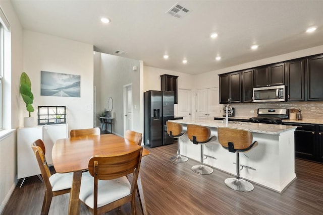 kitchen with appliances with stainless steel finishes, dark wood-type flooring, an island with sink, backsplash, and light stone countertops