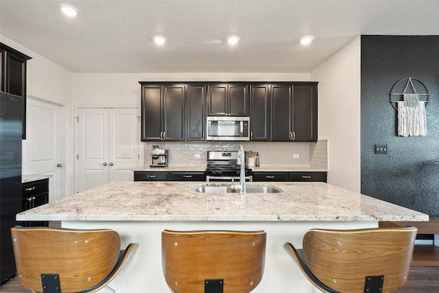 kitchen featuring appliances with stainless steel finishes, tasteful backsplash, a sink, an island with sink, and light stone counters