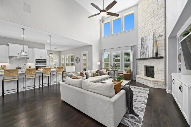 living room featuring a towering ceiling, a stone fireplace, ceiling fan, and dark wood finished floors