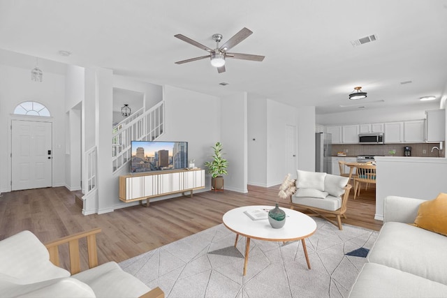 living room featuring stairs, light wood-style floors, ceiling fan, baseboards, and visible vents