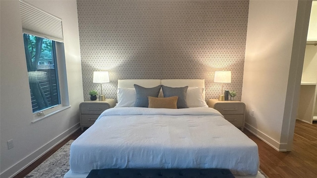bedroom with baseboards, an accent wall, wallpapered walls, and dark wood-style floors