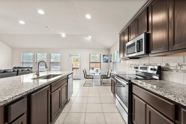 kitchen featuring appliances with stainless steel finishes, a sink, vaulted ceiling, dark brown cabinets, and light stone countertops