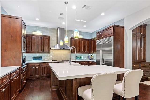 205 Bisset Ct, Austin, TX 78738 - kitchen with built in appliances, wall chimney exhaust hood, light countertops, dark wood finished floors, and a kitchen island with sink