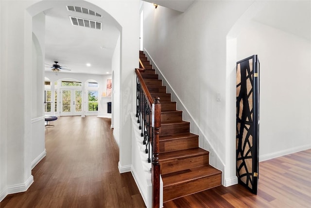 staircase with wood finished floors, arched walkways, ceiling fan, a fireplace with raised hearth, and baseboards