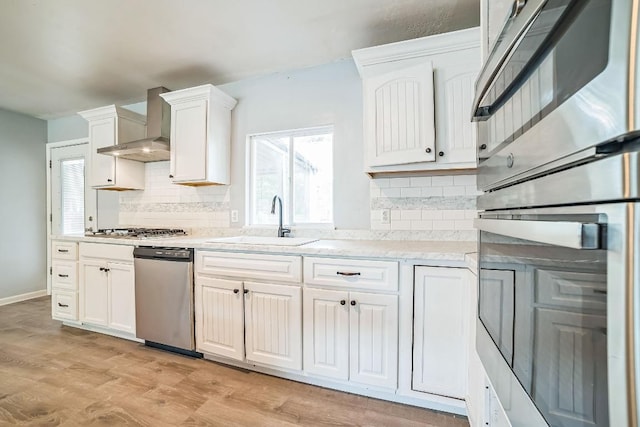kitchen with wall chimney range hood, a sink, white cabinets, stainless steel appliances, and backsplash