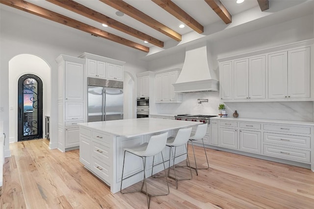 kitchen with a center island, arched walkways, beam ceiling, appliances with stainless steel finishes, and premium range hood