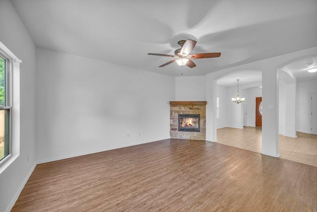 unfurnished living room featuring a stone fireplace, arched walkways, ceiling fan with notable chandelier, and wood finished floors