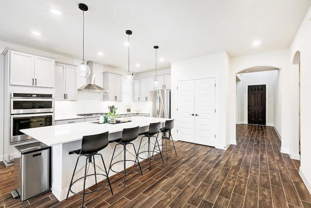 kitchen featuring a kitchen island with sink, dark wood-style flooring, wall chimney range hood, light countertops, and appliances with stainless steel finishes