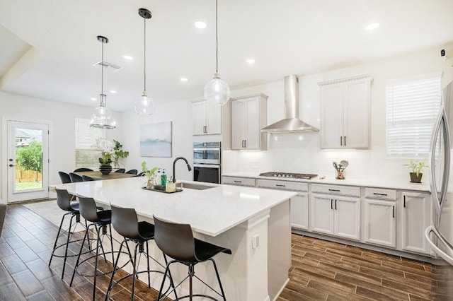 kitchen featuring stainless steel appliances, wood finish floors, wall chimney exhaust hood, a center island with sink, and backsplash