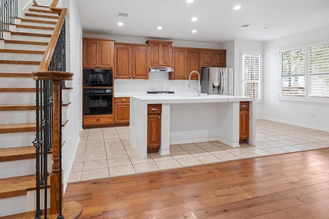 kitchen featuring brown cabinetry, black appliances, under cabinet range hood, and light countertops