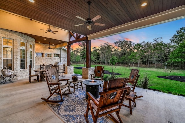 patio terrace at dusk with fence, a lawn, ceiling fan, outdoor dining area, and an outdoor fire pit