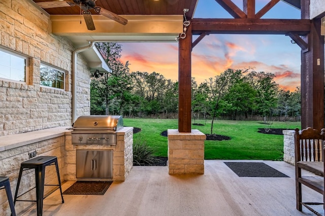 patio terrace at dusk with ceiling fan, an outdoor kitchen, a grill, and a lawn