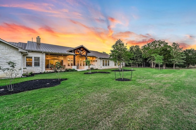 rear view of property with a standing seam roof, stone siding, a chimney, metal roof, and a yard