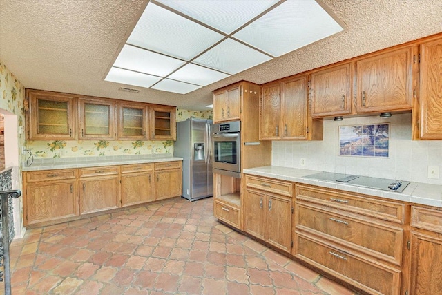 9401 Cedar Crest Dr, Austin, TX 78750 - kitchen featuring stainless steel appliances, decorative backsplash, brown cabinetry, a textured ceiling, and wallpapered walls