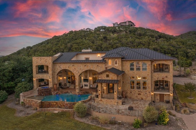 8315 Lime Creek Rd, Volente, TX 78641 - back of house at dusk featuring a standing seam roof, a patio area, stone siding, metal roof, and a wooded view