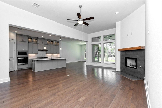 unfurnished living room with a tile fireplace, ceiling fan, visible vents, baseboards, and dark wood-style flooring