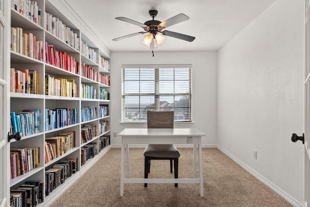 carpeted home office featuring ceiling fan, baseboards, and wall of books