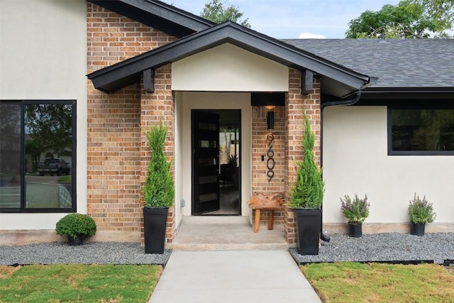 entrance to property featuring a shingled roof, brick siding, and stucco siding