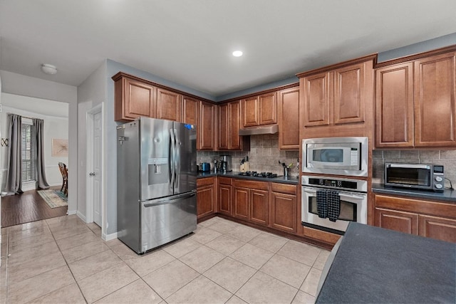 kitchen featuring under cabinet range hood, stainless steel appliances, dark countertops, light tile patterned floors, and decorative backsplash
