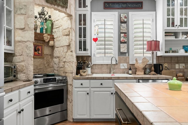 kitchen featuring tile counters, a sink, stainless steel gas range, glass insert cabinets, and white cabinets