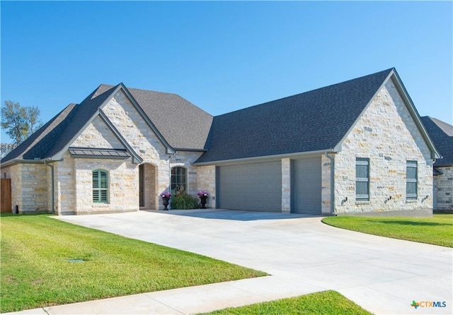 french country inspired facade with concrete driveway, stone siding, an attached garage, a front yard, and roof with shingles