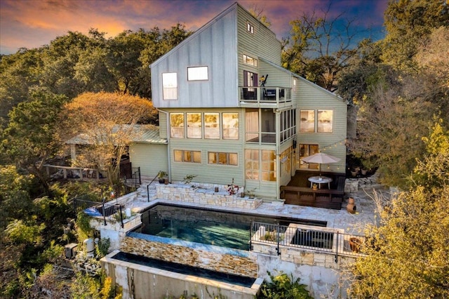 back of house at dusk with board and batten siding, a hot tub, an outdoor pool, and a balcony
