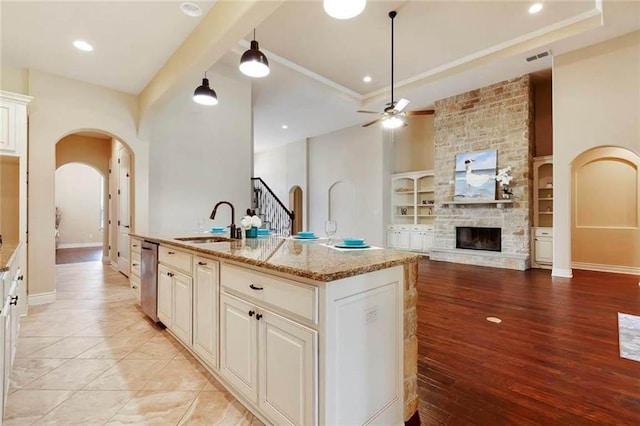 kitchen with ceiling fan, a stone fireplace, arched walkways, a sink, and an island with sink