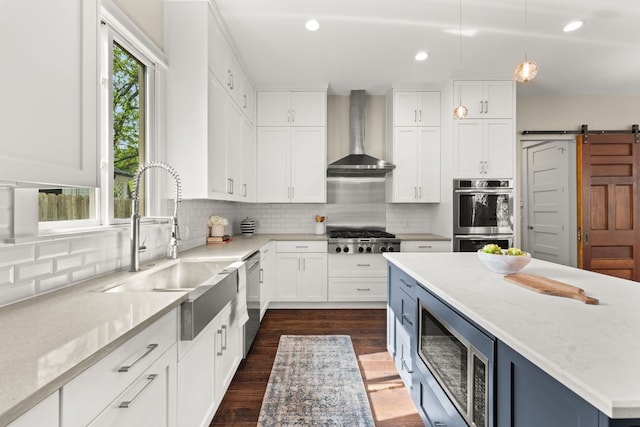 kitchen featuring a barn door, wall chimney range hood, appliances with stainless steel finishes, and white cabinets