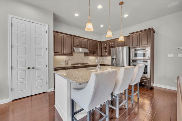 kitchen with under cabinet range hood, stainless steel appliances, backsplash, a sink, and dark wood-style flooring