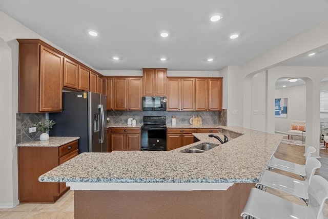 kitchen featuring a sink, arched walkways, black appliances, a kitchen bar, and brown cabinetry