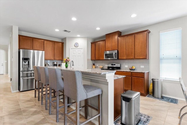30305 Tiger Woods Dr, Georgetown, TX 78628 - kitchen featuring appliances with stainless steel finishes, brown cabinetry, a center island with sink, decorative backsplash, and a breakfast bar area