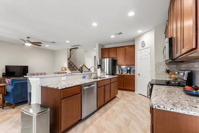 30305 Tiger Woods Dr, Georgetown, TX 78628 - kitchen featuring appliances with stainless steel finishes, a sink, brown cabinets, ceiling fan, and a kitchen island with sink