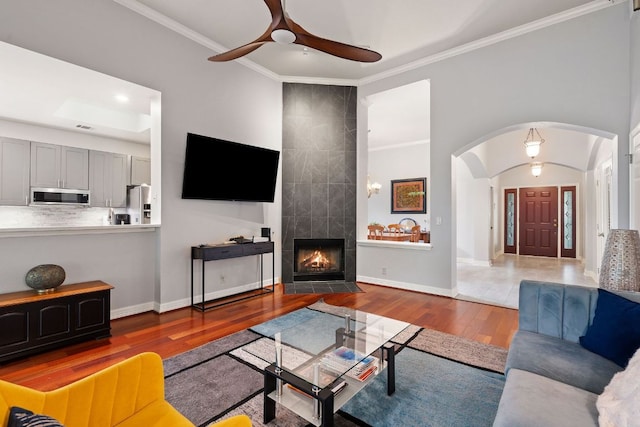 living area with wood finished floors, a ceiling fan, arched walkways, crown molding, and a tiled fireplace