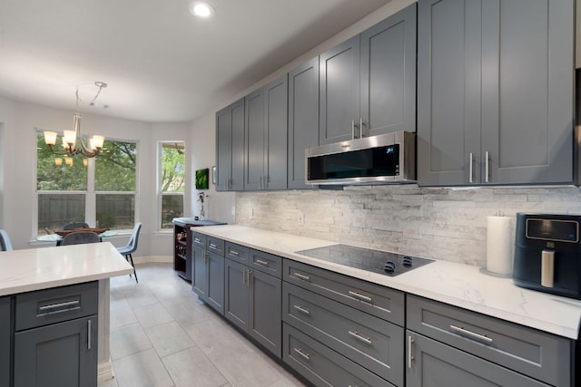 kitchen with backsplash, stainless steel microwave, light stone countertops, black electric stovetop, and a notable chandelier