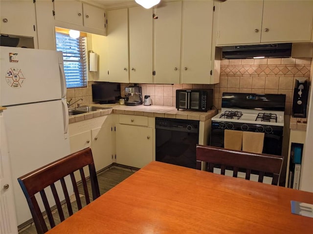 kitchen featuring tile counters, gas stove, under cabinet range hood, black dishwasher, and freestanding refrigerator
