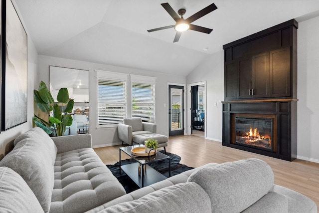 living area with vaulted ceiling, a ceiling fan, a large fireplace, light wood finished floors, and baseboards