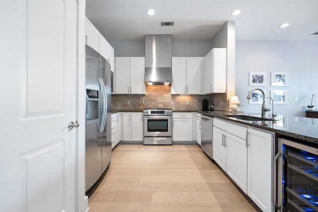 kitchen with beverage cooler, stainless steel appliances, a sink, wall chimney exhaust hood, and dark stone counters