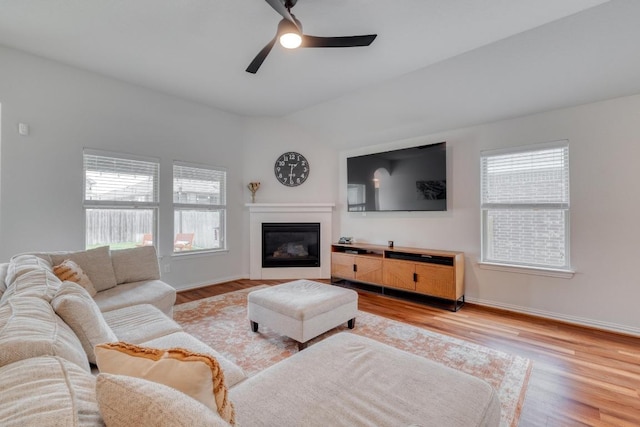 living room with vaulted ceiling, a glass covered fireplace, ceiling fan, light wood finished floors, and baseboards