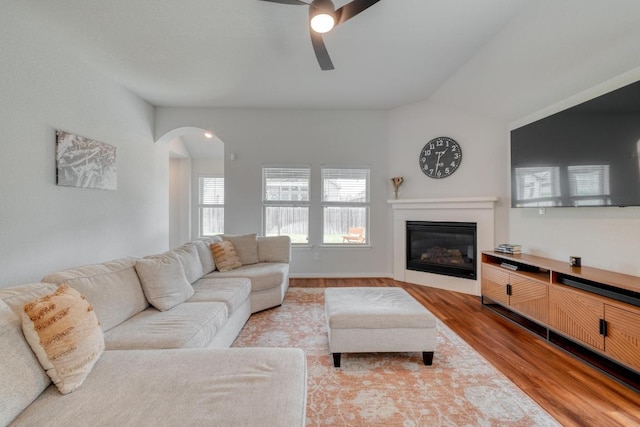 living room featuring vaulted ceiling, arched walkways, light wood-style floors, a glass covered fireplace, and ceiling fan