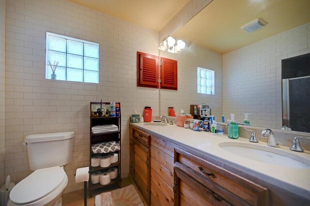 full bath with toilet, a sink, double vanity, and tile walls