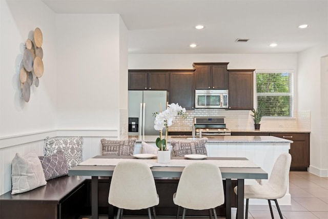 kitchen featuring a kitchen island with sink, dark brown cabinetry, backsplash, light tile patterned floors, and stainless steel appliances