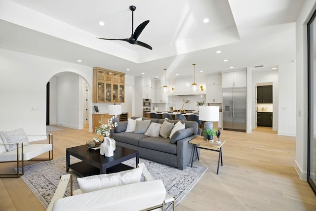 living area with recessed lighting, ceiling fan, a raised ceiling, light wood-style flooring, and arched walkways