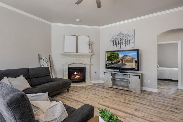 living area featuring crown molding, a warm lit fireplace, ceiling fan, light wood-style flooring, and baseboards