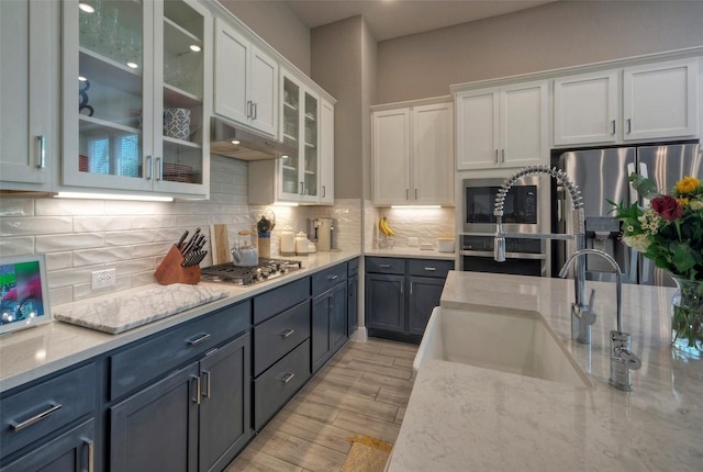 kitchen with light stone countertops, stainless steel appliances, under cabinet range hood, white cabinets, and a sink