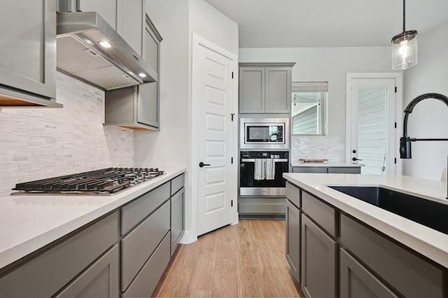kitchen featuring a sink, stainless steel appliances, decorative backsplash, wall chimney exhaust hood, and gray cabinetry