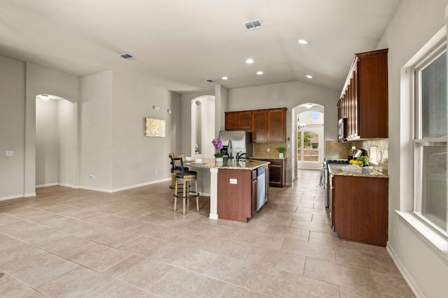 kitchen with a kitchen island with sink, backsplash, vaulted ceiling, arched walkways, and appliances with stainless steel finishes