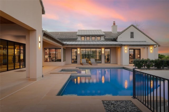 back of house at dusk featuring a standing seam roof, a patio, metal roof, a chimney, and stucco siding