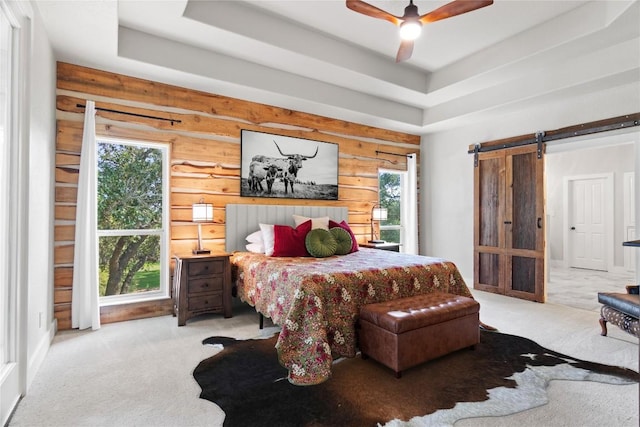 carpeted bedroom with a barn door, a raised ceiling, a ceiling fan, and multiple windows