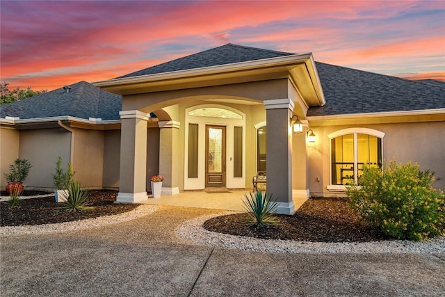 exterior entry at dusk featuring a porch, a shingled roof, and stucco siding