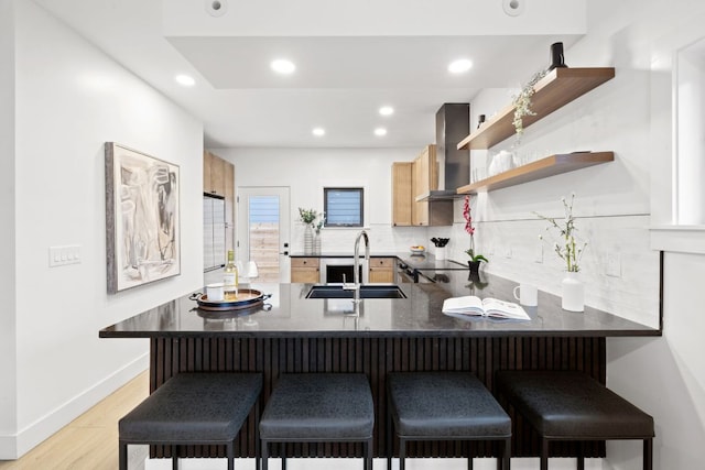 kitchen with a sink, light wood-style flooring, wall chimney exhaust hood, a peninsula, and decorative backsplash